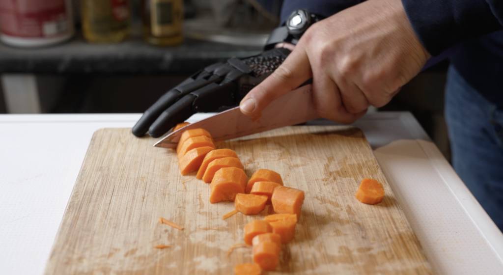 The image shows a close up of Superman Chohan’s hands using a cutting board to slice carrots with a kitchen knife. He is wearing the hero gauntlet on his right forearm and hand, which aids in gripping and controlling the knife. The prosthetic hand appears to be well-integrated into the task, demonstrating its functionality in everyday kitchen activities.