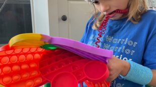 A young child is seated inside, focused on a colorful pop up fidget toy. The child is wearing a blue t-shirt with "Warrior" printed on it and has a complex array of EEG sensors attached to their head. They are sitting in a green chair with a casual home environment around them.