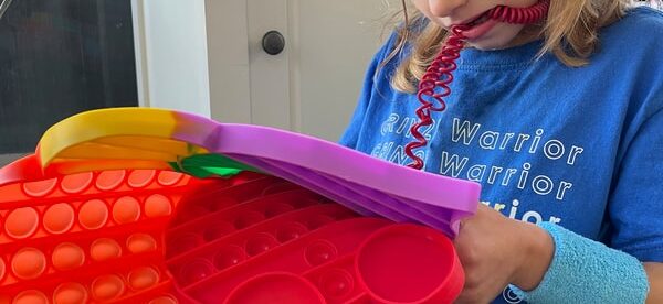 A young child is seated inside, focused on a colorful pop up fidget toy. The child is wearing a blue t-shirt with 