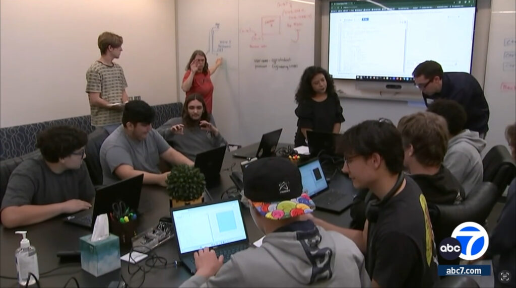 The image shows a group of students, both male and female, participating in a coding bootcamp. They are sitting together around a table with laptops in front of them, working on coding projects or assignments. In the background, there is a whiteboard with diagrams and notes related to coding or programming concepts. The setup suggests an interactive learning environment focused on developing coding skills and understanding programming fundamentals. The students appear engaged and collaborating with one another as they work on their tasks.