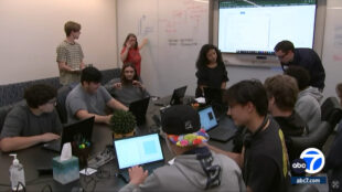 The image shows a group of students, both male and female, participating in a coding bootcamp. They are sitting together around a table with laptops in front of them, working on coding projects or assignments. In the background, there is a whiteboard with diagrams and notes related to coding or programming concepts. The setup suggests an interactive learning environment focused on developing coding skills and understanding programming fundamentals. The students appear engaged and collaborating with one another as they work on their tasks.