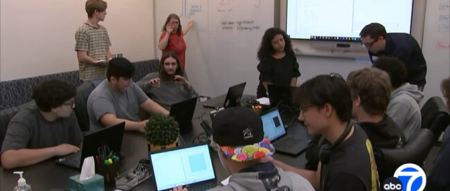 The image shows a group of students, both male and female, participating in a coding bootcamp. They are sitting together around a table with laptops in front of them, working on coding projects or assignments. In the background, there is a whiteboard with diagrams and notes related to coding or programming concepts. The setup suggests an interactive learning environment focused on developing coding skills and understanding programming fundamentals. The students appear engaged and collaborating with one another as they work on their tasks.