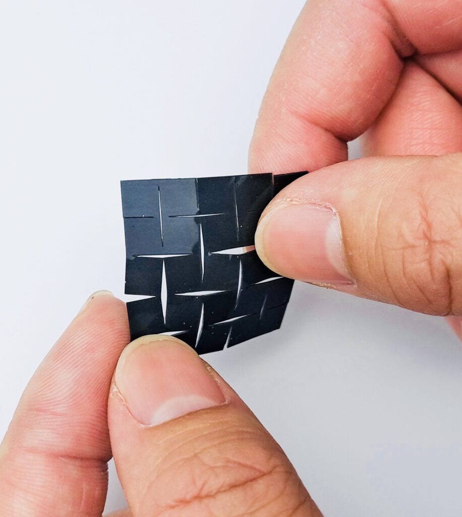 The photo displays a person holding a small, flexible square device between their fingers, showcasing its elasticity and bendable nature. The device appears to be made of a black, grid-like material with cuts that allow it to stretch and conform to different shapes.