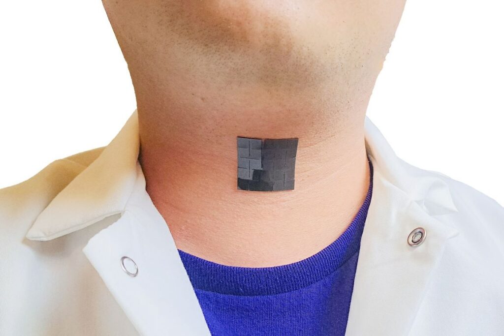 The image shows a person wearing a white shirt and a blue undershirt with a small, black, square bioelectronic device adhered to their neck. The placement on the neck suggests it is positioned over the larynx, aligning with the technology designed to capture muscle movements and translate them into speech.