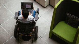 The image shows an overhead view of a person in a motorized wheelchair using a laptop placed on their lap. The person is in an indoor setting with a tiled floor. To the right, there is a modern, green armchair with a patterned cushion. The space appears to be a public area, possibly a lobby or a lounge.