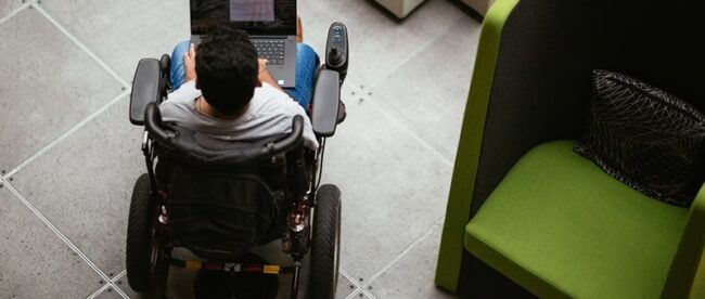 The image shows an overhead view of a person in a motorized wheelchair using a laptop placed on their lap. The person is in an indoor setting with a tiled floor. To the right, there is a modern, green armchair with a patterned cushion. The space appears to be a public area, possibly a lobby or a lounge.