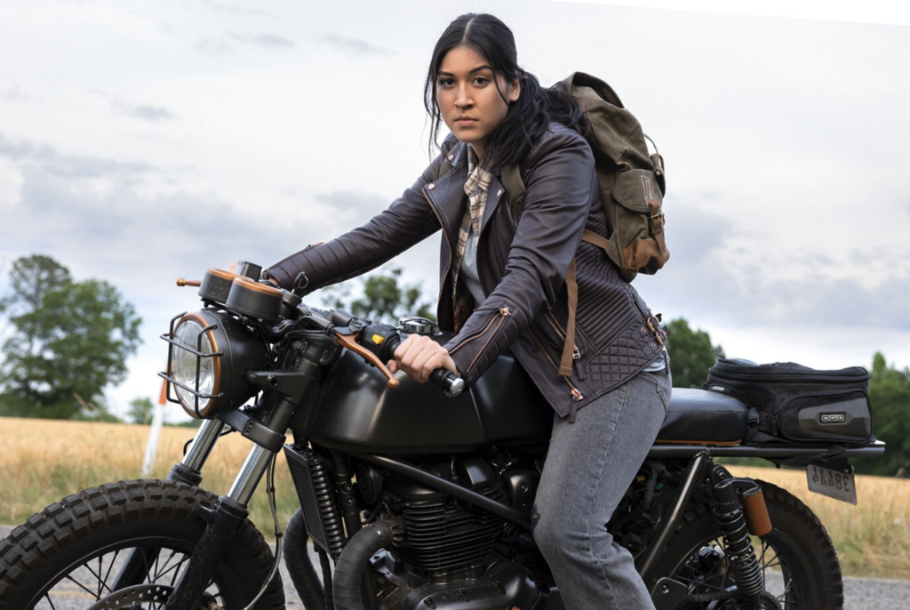 Alaqua Cox is seated on a black Royal Enfield Continental GT 650 motorcycle, parked outdoors with fields in the background and is looking straight into the camera. She is wearing a brown leather jacket, a plaid shirt, dark pants, and has a backpack on.