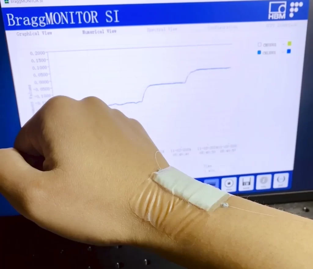 The image shows a person's hand wearing a wearable sensor device on their wrist, with the sensor connected to a computer screen displaying graphs and data related to monitoring or tracking movements or signals from the wearable sensor. The software on the screen is titled "BraggMONITOR SI" and appears to be used for analyzing data from fiber optic sensors that detect bending or motion.