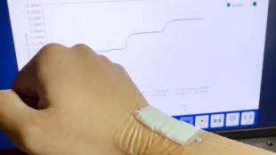 The image shows a person's hand wearing a wearable sensor device on their wrist, with the sensor connected to a computer screen displaying graphs and data related to monitoring or tracking movements or signals from the wearable sensor. The software on the screen is titled "BraggMONITOR SI" and appears to be used for analyzing data from fiber optic sensors that detect bending or motion.