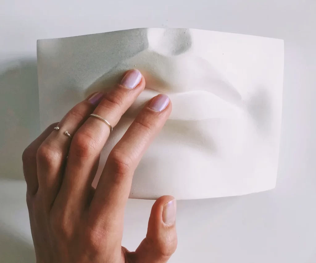 The image features a hand interacting with a tactile piece of artwork. The hand is shown with the fingers gently pressing against a flexible, translucent material with an embossed or raised design resembling facial features. The thumb and forefinger are in focus, touching the material, which seems to capture the contours of a human face in a three-dimensional form. The artwork appears to be designed to convey visual details through touch, likely for a sensory or educational purpose. The person's nails are neatly manicured with a subtle polish, and they wear a simple ring on the ring fingering middle finger.