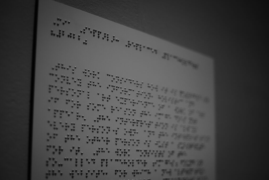 The image depicts a close-up of a board with text written in Braille. The raised dots are arranged in the distinctive patterns that represent letters and numbers in the Braille system, which is designed for tactile reading by individuals who are blind or have low vision. The focus and lighting of the photograph emphasize the tactile nature of the Braille, with shadows accentuating the three-dimensional aspect of the dots. The monochrome color scheme adds to the image's texture-focused aesthetic.