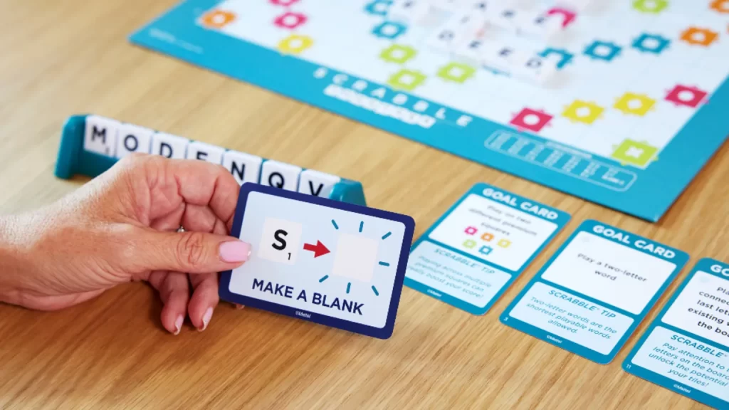 a close-up of a hand holding a Scrabble game card with instructions to "MAKE A BLANK" alongside a letter tile rack with the letters "MODENQV". The background reveals a part of the Scrabble Together game, suggesting an ongoing game with a focus on the card and the player's hand.