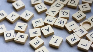 scattered arrangement of Scrabble letter tiles face up on a grey background. The tiles are in disarray, providing a sense of the variety and randomness of letters available in the game. There is a blank tile visible among the letters, highlighting the potential for creating any letter a player needs.