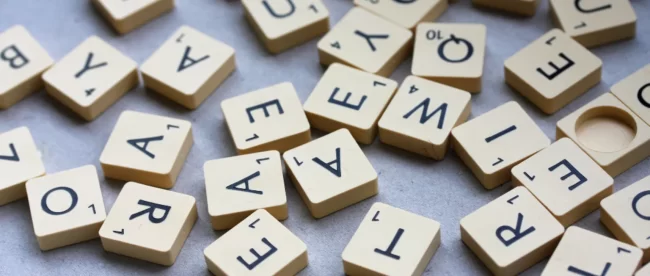 scattered arrangement of Scrabble letter tiles face up on a grey background. The tiles are in disarray, providing a sense of the variety and randomness of letters available in the game. There is a blank tile visible among the letters, highlighting the potential for creating any letter a player needs.