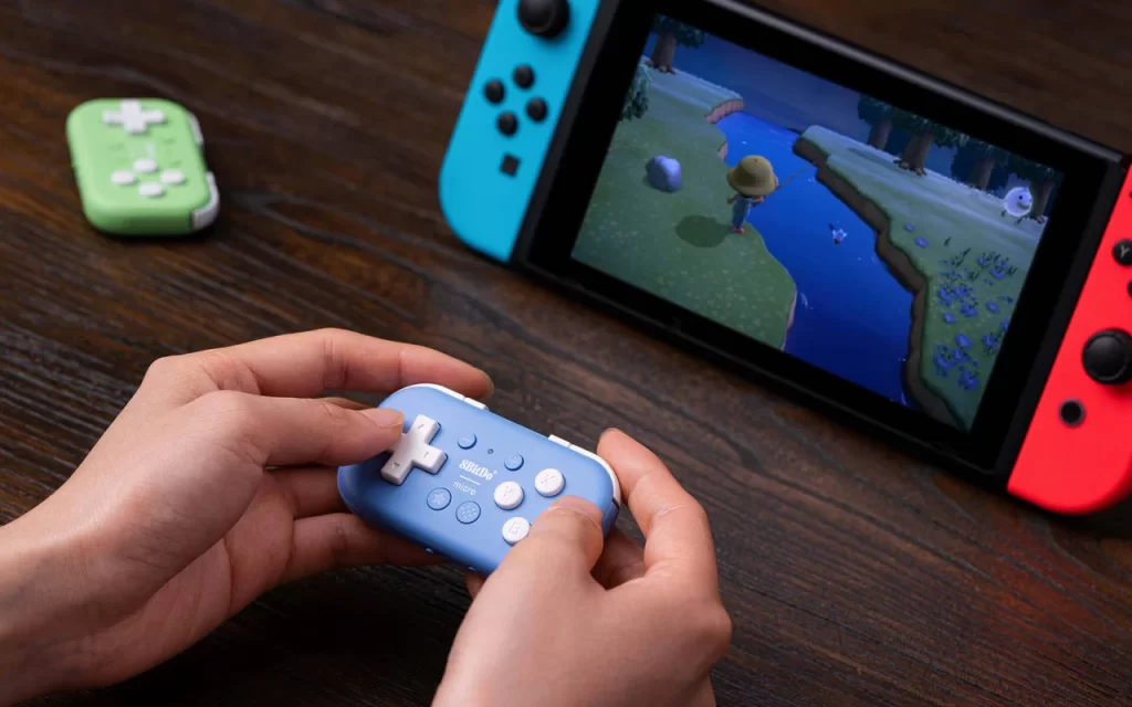 image shows a person playing a game on a Nintendo Switch, which is placed in its handheld docking mode with Joy-Con controllers attached in blue and red. The player is using a compact, light blue 8BitDo Micro Bluetooth gamepad to control the game, which appears to be "Animal Crossing: New Horizons".