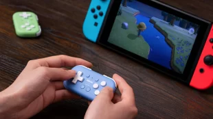 image shows a person playing a game on a Nintendo Switch, which is placed in its handheld docking mode with Joy-Con controllers attached in blue and red. The player is using a compact, light blue 8BitDo Micro Bluetooth gamepad to control the game, which appears to be "Animal Crossing: New Horizons".