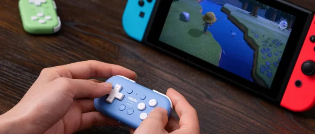 image shows a person playing a game on a Nintendo Switch, which is placed in its handheld docking mode with Joy-Con controllers attached in blue and red. The player is using a compact, light blue 8BitDo Micro Bluetooth gamepad to control the game, which appears to be 