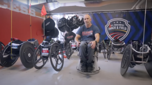 Adam Bleakney is seated in the PURE hands-free wheelchair prototype in a room filled with various racing wheelchairs and equipment. The backdrop features a U.S. Paralympics Track & Field banner, emphasizing the connection to athleticism and innovation in wheelchair design.