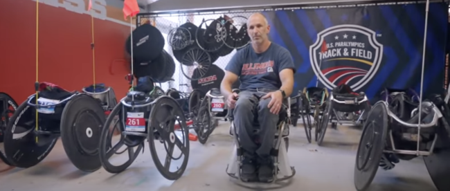 Adam Bleakney is seated in the PURE hands-free wheelchair prototype in a room filled with various racing wheelchairs and equipment. The backdrop features a U.S. Paralympics Track & Field banner, emphasizing the connection to athleticism and innovation in wheelchair design.