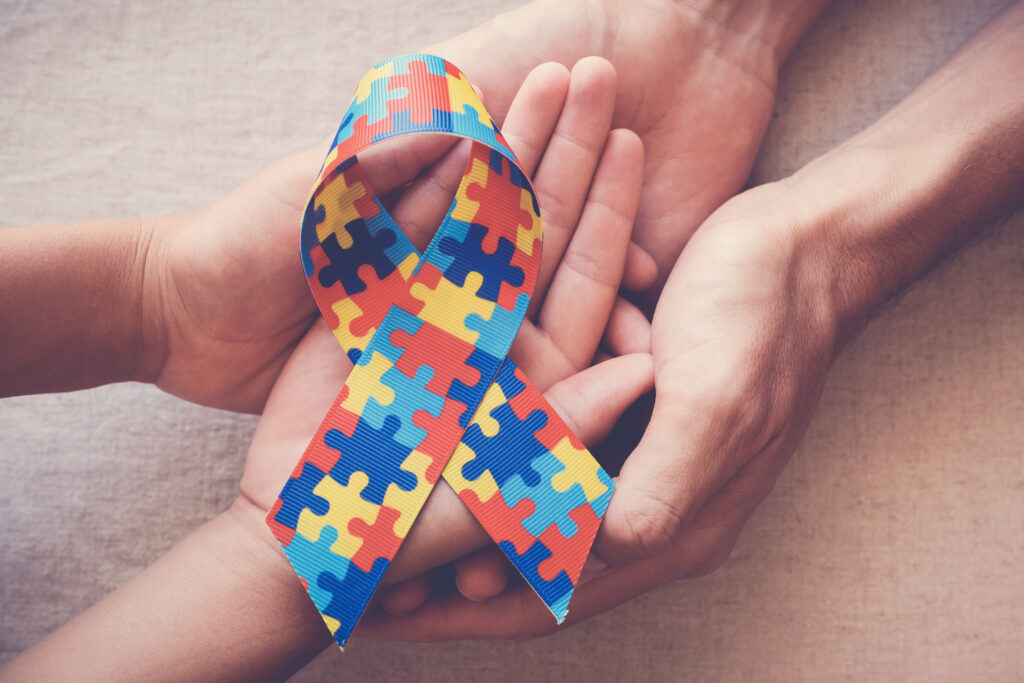 The image shows a multicolored puzzle ribbon, which is commonly associated with autism awareness. The ribbon is held by multiple hands, symbolizing support and unity in the autism community. The hands appear to be of different ages, suggesting a connection between children and adults in the effort to raise awareness and provide support for autism.