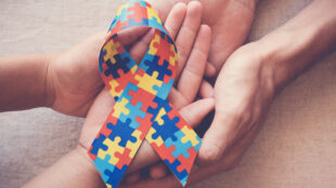 The image shows a multicolored puzzle ribbon, which is commonly associated with autism awareness. The ribbon is held by multiple hands, symbolizing support and unity in the autism community. The hands appear to be of different ages, suggesting a connection between children and adults in the effort to raise awareness and provide support for autism.