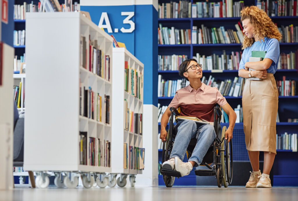 A person using a wheelchair is smiling and engaging in a conversation with a standing individual in a library setting. They are surrounded by bookshelves filled with books, creating a vibrant and inclusive atmosphere. Both individuals appear to be in a positive and friendly interaction.