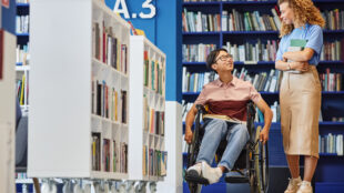 A person using a wheelchair is smiling and engaging in a conversation with a standing individual in a library setting. They are surrounded by bookshelves filled with books, creating a vibrant and inclusive atmosphere. Both individuals appear to be in a positive and friendly interaction.