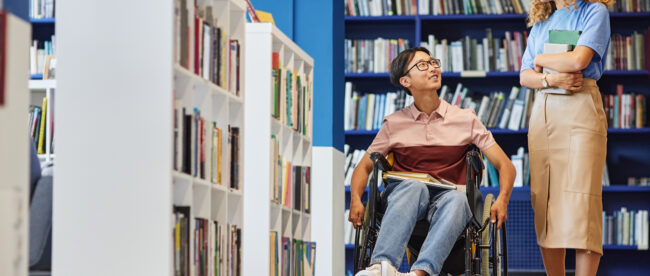 A person using a wheelchair is smiling and engaging in a conversation with a standing individual in a library setting. They are surrounded by bookshelves filled with books, creating a vibrant and inclusive atmosphere. Both individuals appear to be in a positive and friendly interaction.