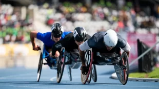 Three athletes in specialized racing wheelchairs compete intensely in a track event. The athlete in the foreground wears a white helmet and a black racing suit, leaning forward with determination. Right behind him is another athlete in a black helmet and suit. The third athlete, wears a black helmet and a blue racing suit. All three athletes are pushing the rims of their racing wheelchairs with their hands. The blurred background reveals a stadium filled with spectators, creating a dynamic and energetic atmosphere.