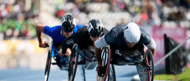 Three athletes in specialized racing wheelchairs compete intensely in a track event. The athlete in the foreground wears a white helmet and a black racing suit, leaning forward with determination. Right behind him is another athlete in a black helmet and suit. The third athlete, wears a black helmet and a blue racing suit. All three athletes are pushing the rims of their racing wheelchairs with their hands. The blurred background reveals a stadium filled with spectators, creating a dynamic and energetic atmosphere.