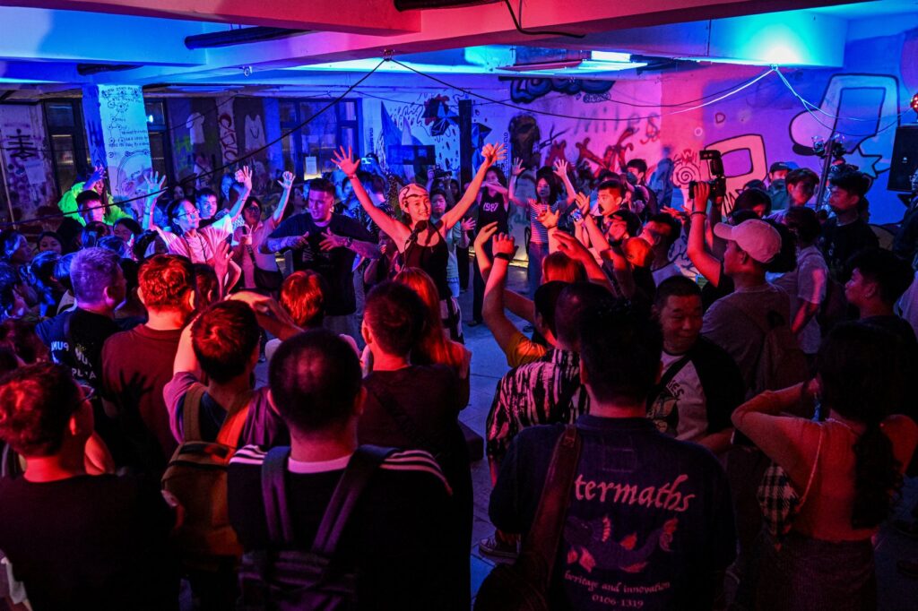 The image depicts a vibrant scene at a nightclub where a large crowd of people, both deaf and hearing, are enthusiastically dancing and raising their hands. The room is bathed in colorful lights, with shades of blue, pink, and purple illuminating the space. The diverse group of attendees appears fully engaged, enjoying the music and the inclusive atmosphere. The graffiti-covered walls in the background add to the energetic and urban vibe of the event.