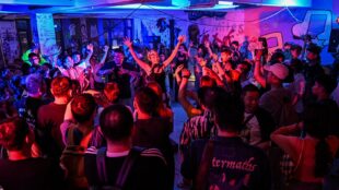 The image depicts a vibrant scene at a nightclub where a large crowd of people, both deaf and hearing, are enthusiastically dancing and raising their hands. The room is bathed in colorful lights, with shades of blue, pink, and purple illuminating the space. The diverse group of attendees appears fully engaged, enjoying the music and the inclusive atmosphere. The graffiti-covered walls in the background add to the energetic and urban vibe of the event.