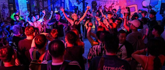 The image depicts a vibrant scene at a nightclub where a large crowd of people, both deaf and hearing, are enthusiastically dancing and raising their hands. The room is bathed in colorful lights, with shades of blue, pink, and purple illuminating the space. The diverse group of attendees appears fully engaged, enjoying the music and the inclusive atmosphere. The graffiti-covered walls in the background add to the energetic and urban vibe of the event.