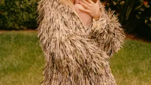 This image features the August 2024 cover of British Vogue, showcasing Suki Waterhouse holding her newborn daughter in a tender embrace. Suki is dressed in a textured, fringe-adorned coat while standing in a lush garden, creating a serene and intimate atmosphere. The cover story, titled "Summer of Love," reflects on Suki’s journey into motherhood, celebrating her new chapter in life with a sense of peace and fulfillment.