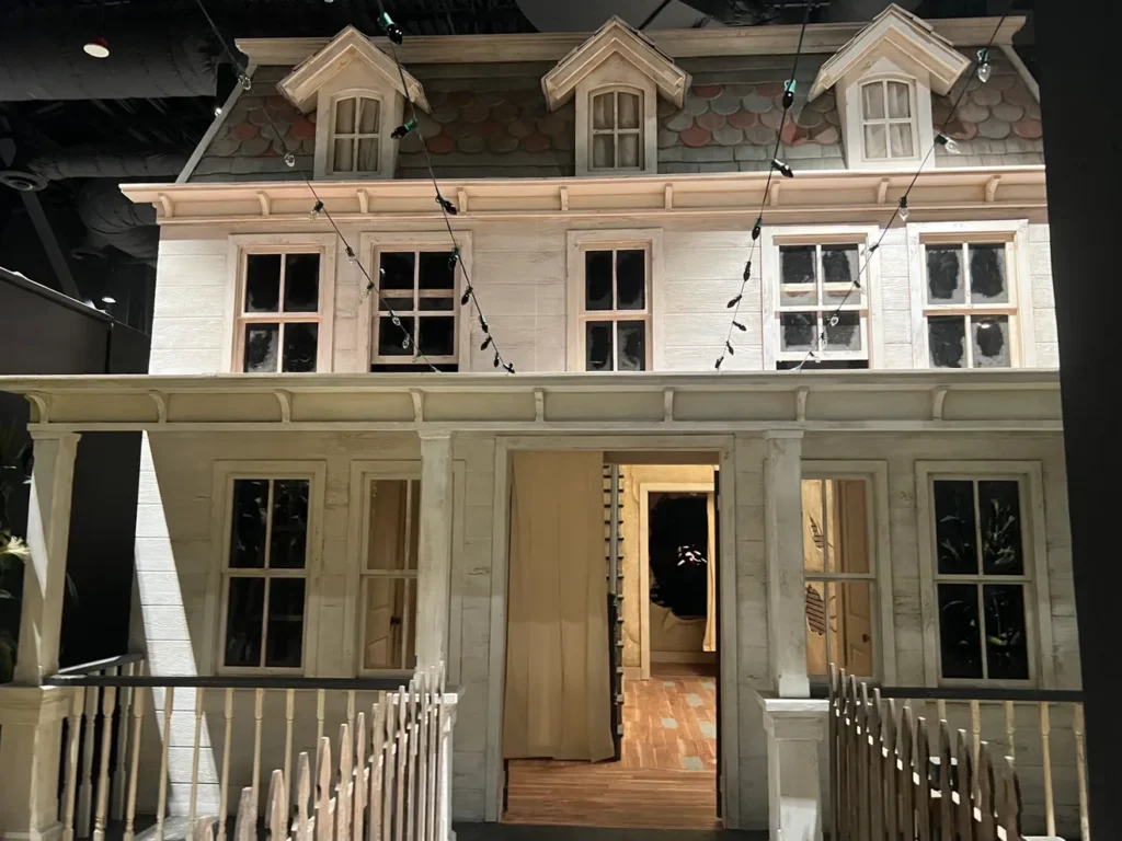 The Abbotts' home from "A Quiet Place" is recreated inside the Universal Studios Hollywood Halloween Horror Nights house of the same name. It's a large, two-story white house facade. The house features a Victorian-style architecture with dormer windows on the upper floor, a wrap-around porch, and detailed trim work. The front door is open, revealing a lit interior. String lights are hung above, and a white picket fence is visible in the foreground. The house appears to be inside a larger indoor space, suggesting it's a prop or attraction.