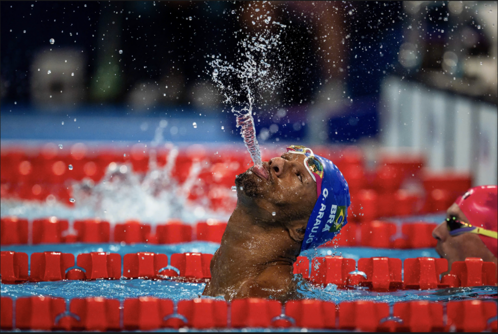 A Brazilian Paralympic swimmer, Gabriel Geraldo dos Santos Araújo, celebrates in the pool by spitting a stream of water high into the air. He wears a blue swim cap with the Brazilian flag and "BRA" visible. The swimmer is surrounded by red lane dividers, and water droplets are scattered in the air around him. His head is tilted back in celebration after winning the men's 100m backstroke S2 final.