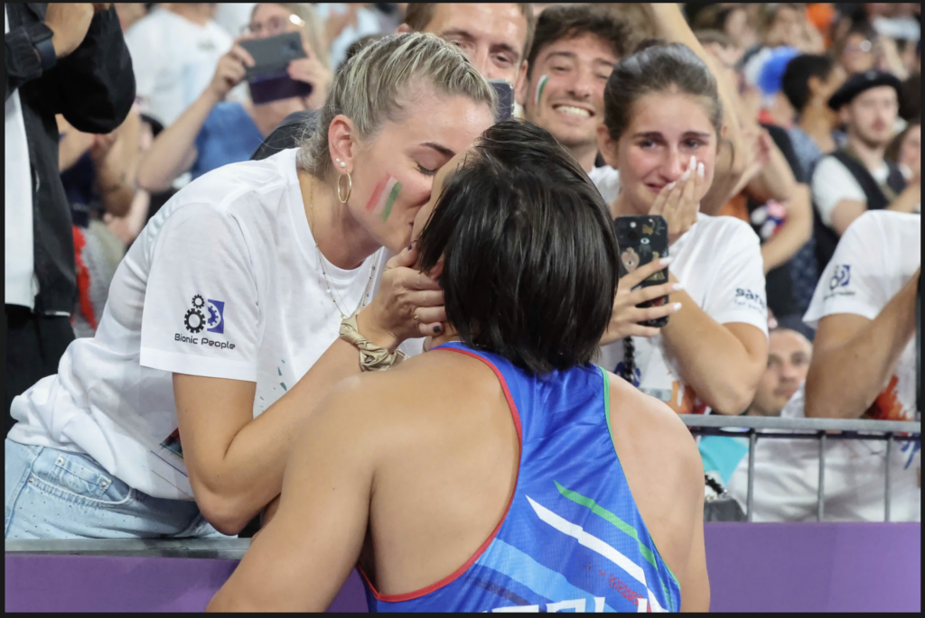 An emotional scene at a sporting event shows a woman in a white "Bionic People" t-shirt leaning over a barrier to kiss an athlete in a blue uniform. The crowd in the background is cheering and taking photos, with one person visibly crying tears of joy.