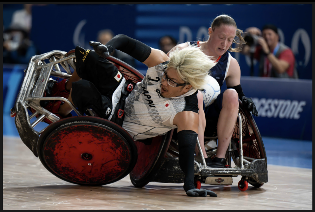 Two wheelchair rugby players collide during an intense match. A Japanese player in a white jersey with blonde hair and glasses is tipping over in their specialized rugby wheelchair. An opponent in a dark jersey is pushing against them. The red wheels of the wheelchair and the wooden court are visible in the foreground.