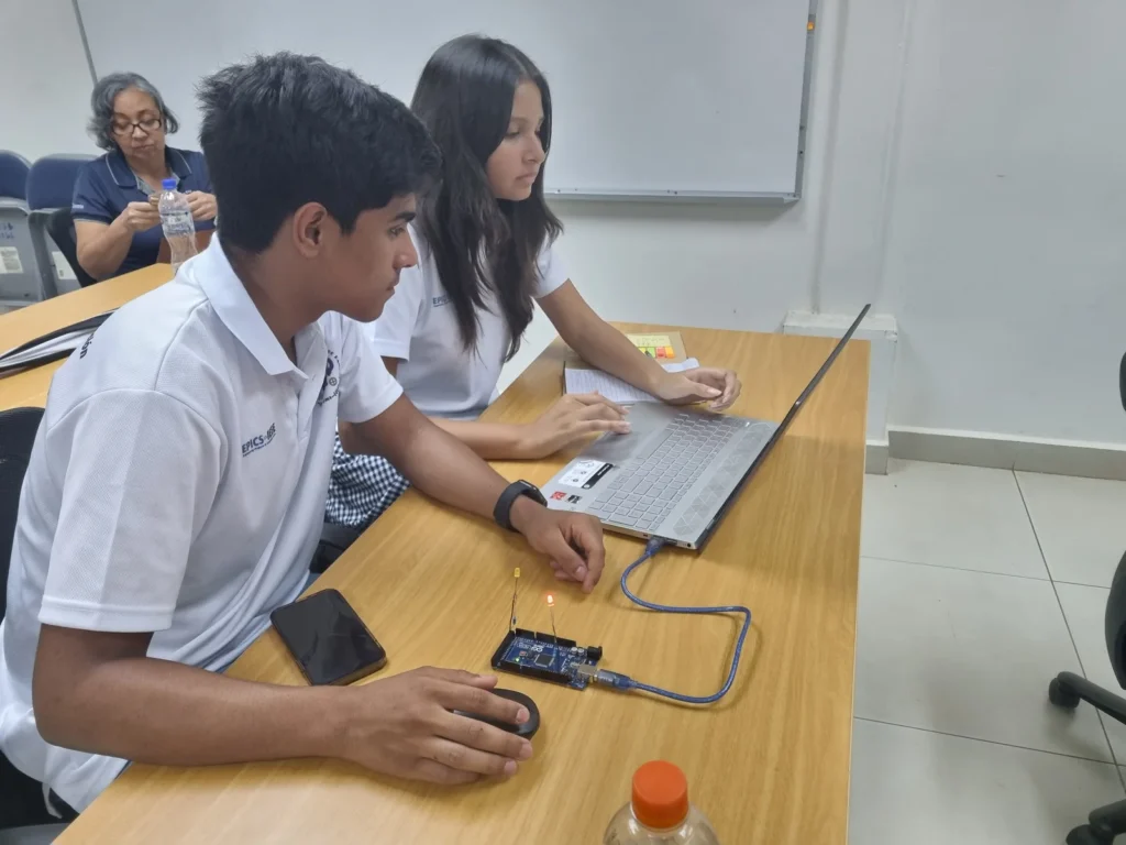 Two students from Universidad Tecnológica de Panamá are seated at a desk, testing a microcontroller-based prototype. One student is using a laptop while the other operates a small electronic circuit connected to the laptop. The circuit is equipped with visible LEDs and wiring, indicating an active project. The students are working collaboratively, on a project aimed at improving accessibility within their school environment. A third person in the background is seated, looking at her phone.