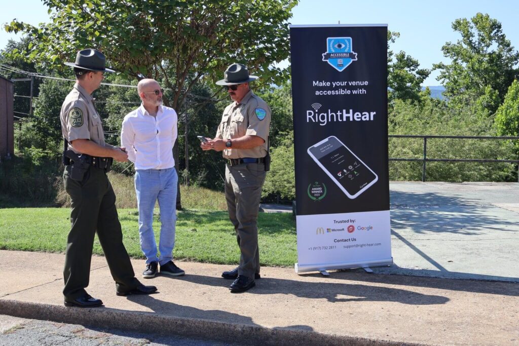 two park rangers and a man in business attire standing next to a promotional banner for RightHear at Natural Bridge State Park, an accessibility solution. The banner highlights how venues can be made accessible using RightHear’s technology, featuring a phone interface. The men appear to be discussing the application, emphasizing efforts to improve accessibility in public spaces. The backdrop includes trees and a sunny outdoor environment, enhancing the image’s focus on accessibility in natural settings.