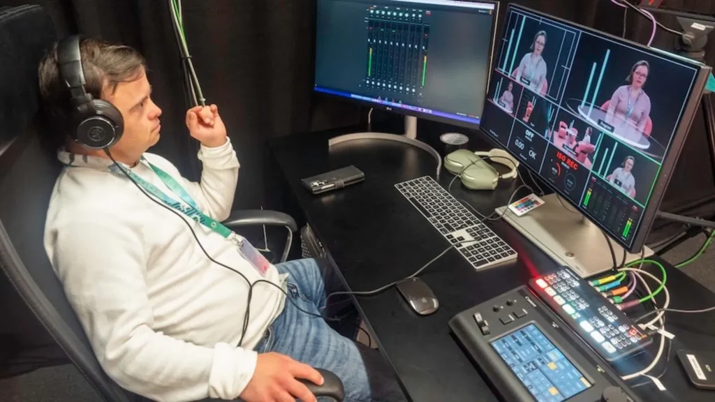 Reporter Svein Andre Hofsø sits watching screens in a television control room. He looks relaxed as he leans back but focused on what he is watching. He is dressed in a light, long-sleeved t-shirt and denim jeans, and wearing headphones and an identity card on a green lanyard. 
