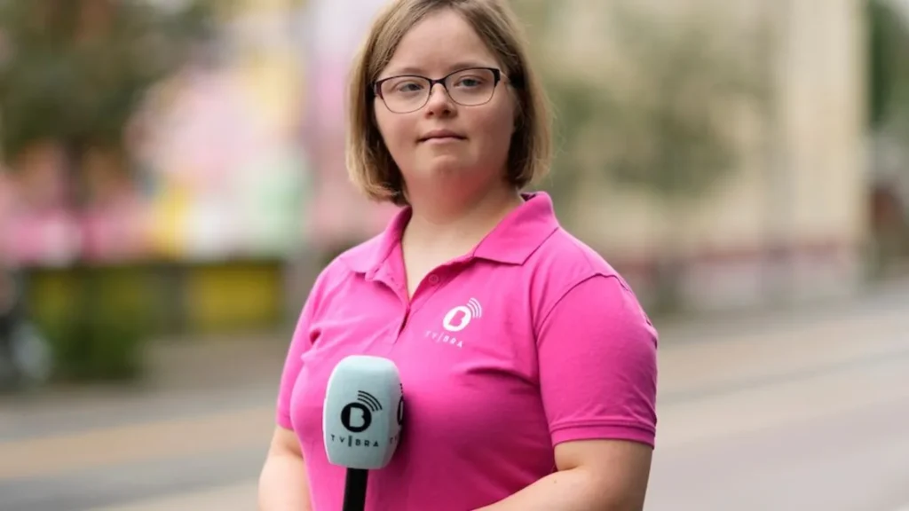 Emily Ann Riedel with Down's syndrome stands in a street, holding a microphone. She is dressed in a bright pink polo shirt. Her hair is medium brown and not quite shoulder length. She is wearing glasses and has a hint of a smile.