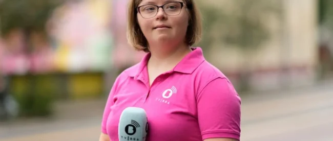 Emily Ann Riedel with Down's syndrome stands in a street, holding a microphone. She is dressed in a bright pink polo shirt. Her hair is medium brown and not quite shoulder length. She is wearing glasses and has a hint of a smile.