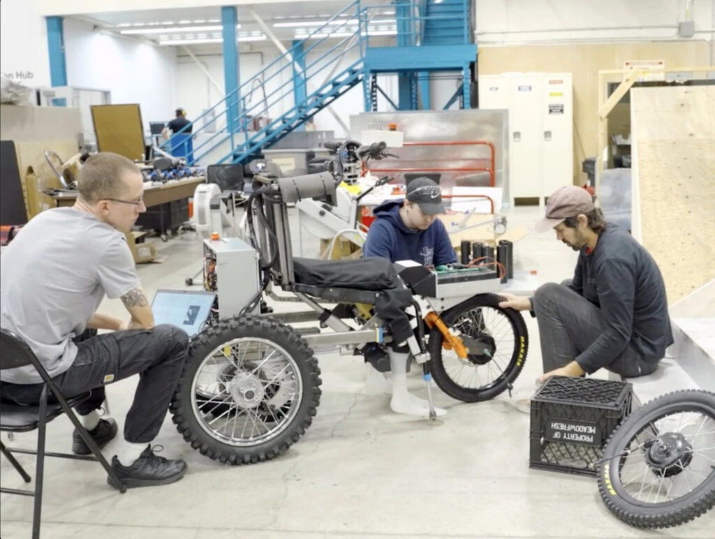 Three men work on an innovative off-road wheelchair in a workshop. The wheelchair has large, rugged tires and a complex frame. One man sits at a laptop, another examines the wheelchair's frame, and the third adjusts the front wheel. The background shows a industrial workspace with stairs and various equipment.​​​​​​​​​​​​​​​​