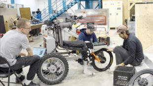 Three men work on an innovative off-road wheelchair in a workshop. The wheelchair has large, rugged tires and a complex frame. One man sits at a laptop, another examines the wheelchair's frame, and the third adjusts the front wheel. The background shows a industrial workspace with stairs and various equipment.​​​​​​​​​​​​​​​​