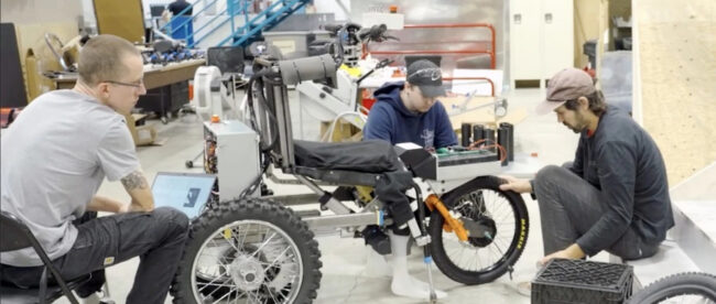 Three men work on an innovative off-road wheelchair in a workshop. The wheelchair has large, rugged tires and a complex frame. One man sits at a laptop, another examines the wheelchair's frame, and the third adjusts the front wheel. The background shows a industrial workspace with stairs and various equipment.​​​​​​​​​​​​​​​​