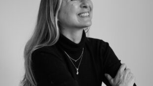 Black & while portrait of Nicky Weir - makeup artist and second guest on the. Shaping Tomorrow podcast. Nicky is seen wearing a black full sleeves shirt, posing at an angle and smiling.