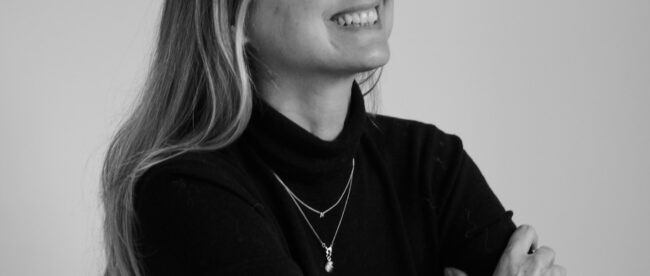 Black & while portrait of Nicky Weir - makeup artist and second guest on the. Shaping Tomorrow podcast. Nicky is seen wearing a black full sleeves shirt, posing at an angle and smiling.