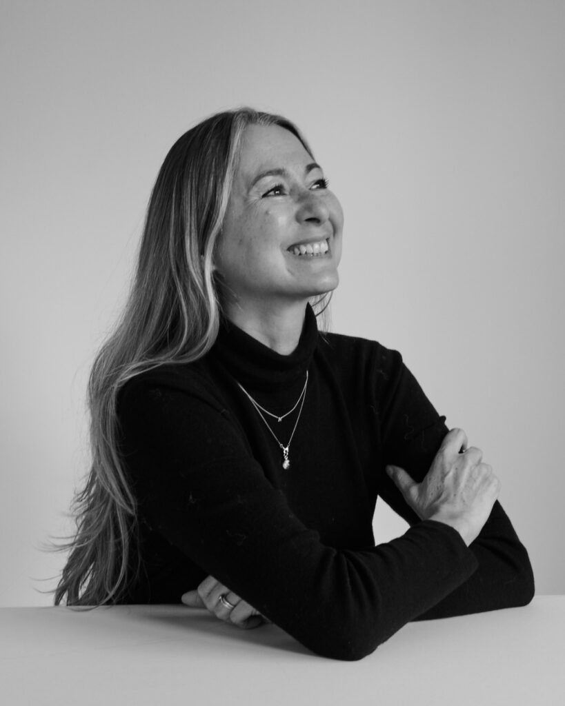 Black & while portrait of Nicky Weir - makeup artist and second guest on the. Shaping Tomorrow podcast. Nicky is seen wearing a black full sleeves shirt, posing at an angle and smiling.