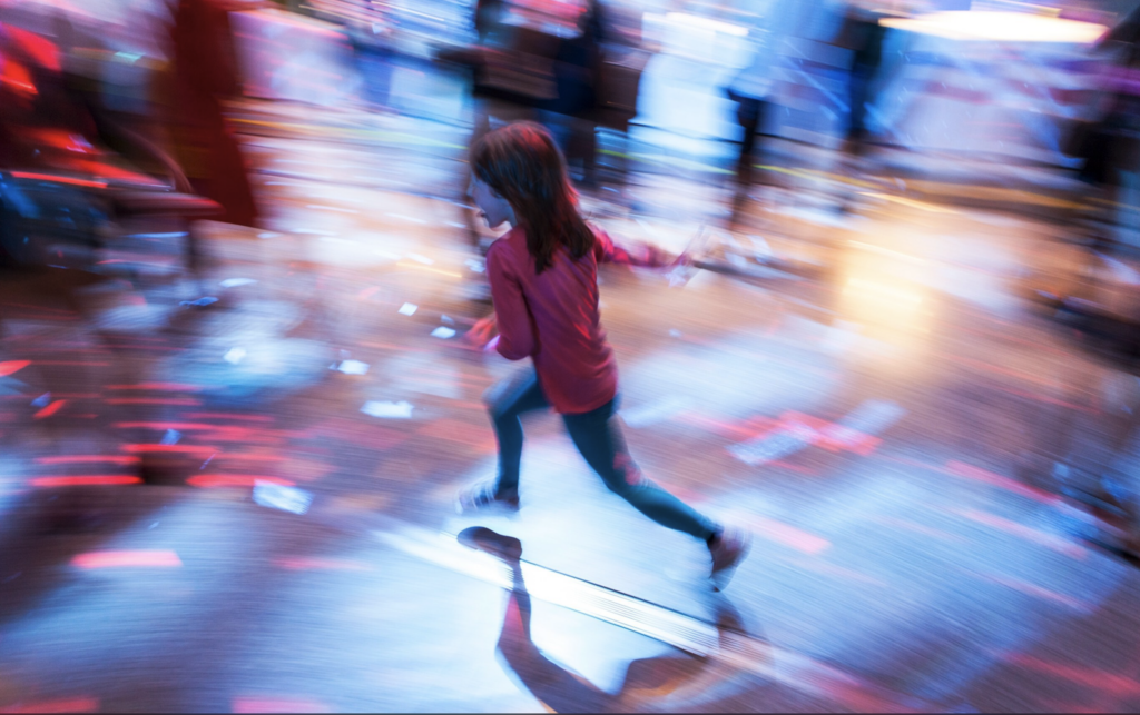 A blurred motion photograph shows a person in a red top and dark pants running or moving quickly across a reflective floor, creating a dynamic sense of movement with blue and red color streaks in the background.